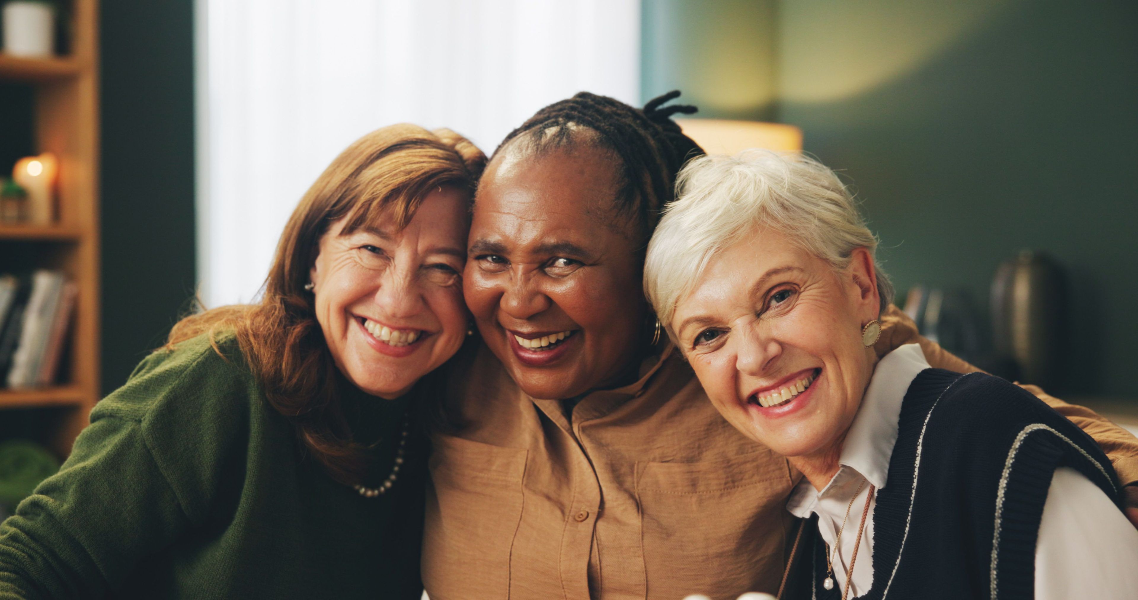 Three diverse senior women smiling warmly and embracing, celebrating friendship and community in a cozy indoor setting.