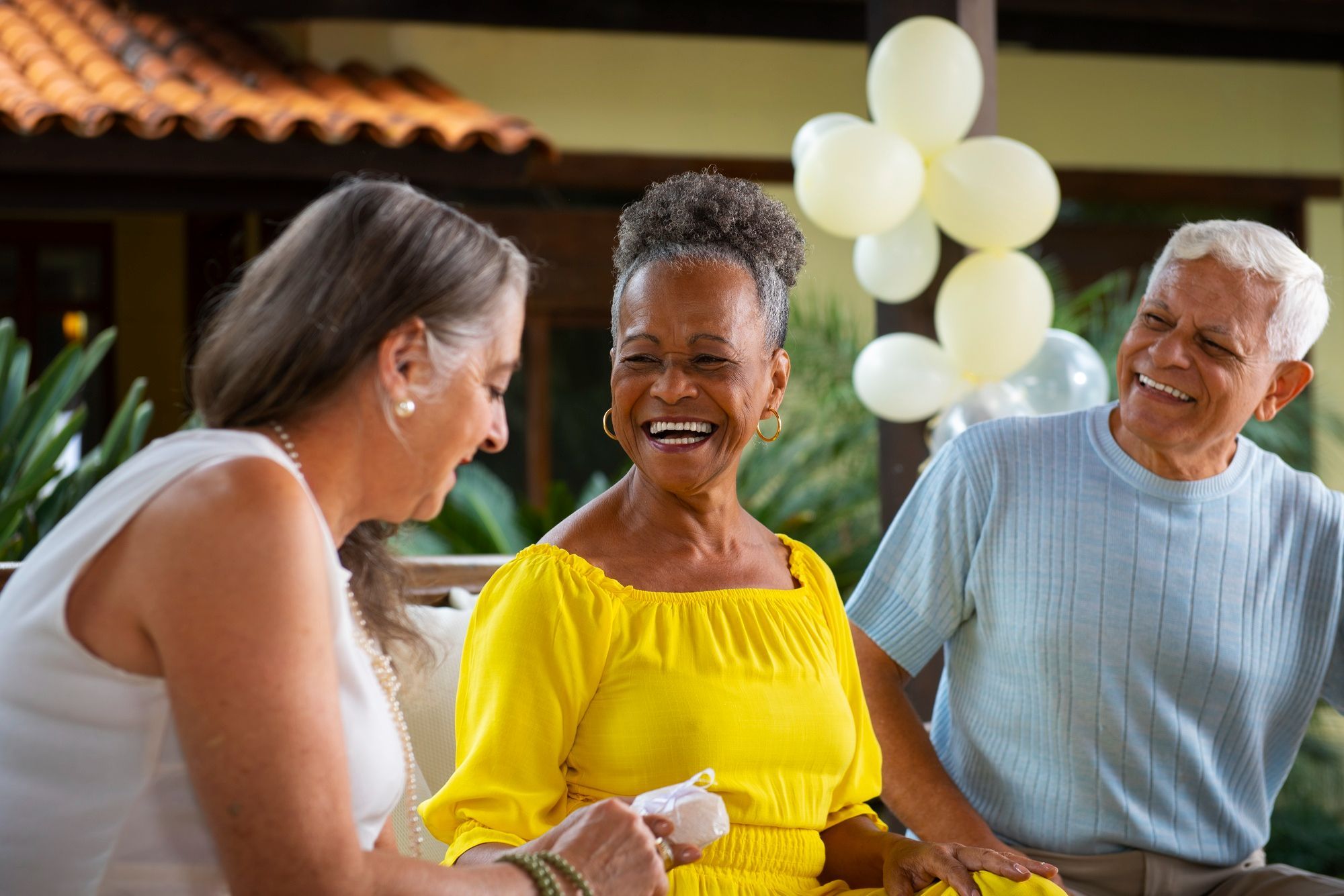 Three seniors laughing together at an outdoor celebration with balloons, enjoying a happy and festive moment.