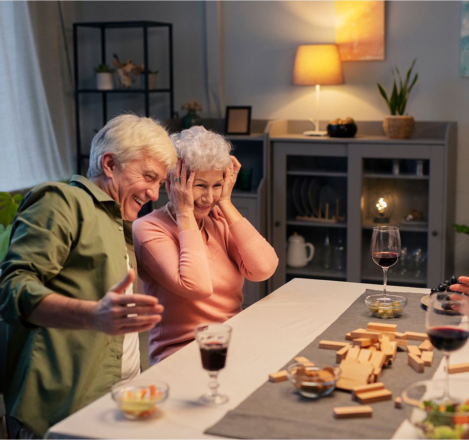 Elderly couple laughing at a game night with wine, snacks, and toppled Jenga pieces, enjoying a playful evening together.