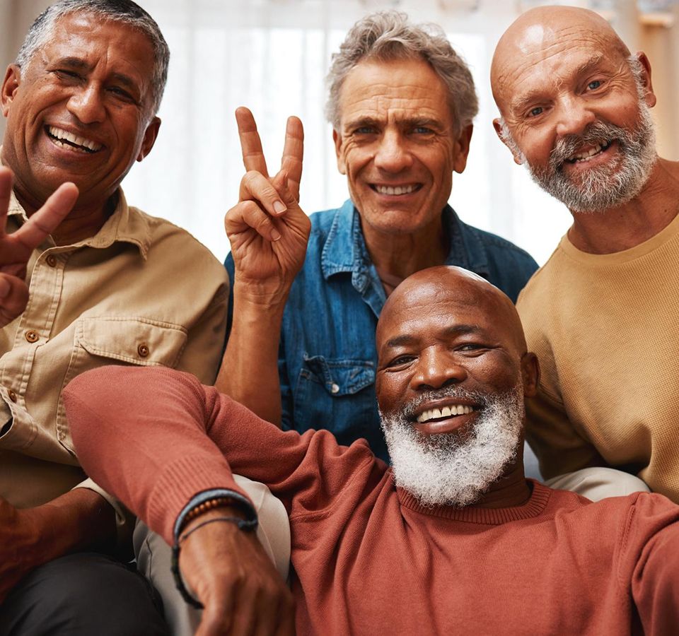Four senior men smiling and flashing peace signs, sharing joy and friendship in a warm, relaxed setting.