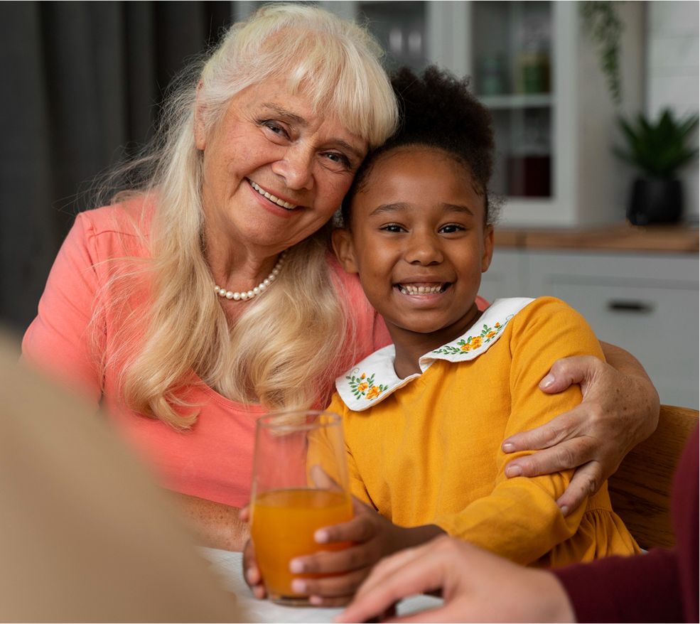 Smiling senior woman sitting with a young girl at a table, holding orange juice and sharing a happy moment.